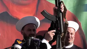 در این تصویر، دو فرد که در حال سخنرانی هستند، به نظر می‌رسد در یک مراسم یا تجمع حضور دارند. یکی از آن‌ها سلاحی را در دست دارد و دیگر فرد در سمت راست نیز در حال نظارت یا همراهی با او است. پس‌زمینه تصویر شامل رنگ‌های زنده و گرافیکی است. هر دو فرد کلاه‌های مخصوص خود را بر سر دارند که نشان‌دهنده هویت مذهبی یا فرهنگی آن‌ها است. احساس کلی تصویر به نظر می‌رسد جدی و تأکیدی بر موضوعی مهم باشد.