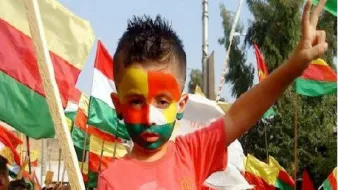 L'image montre un jeune garçon au milieu d'une manifestation ou d'une célébration. Il a le visage peint aux couleurs du drapeau, probablement inspirées par les couleurs du Cameroun, avec des nuances de vert, rouge et jaune. Il porte un t-shirt rouge et affiche un signe de paix avec sa main, tout en étant entouré de drapeaux aux couleurs similaires. L'atmosphère semble festive et engagée.