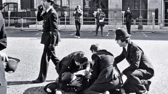 L'image montre une scène policière en noir et blanc. Plusieurs policiers en uniforme entourent une personne au sol, visiblement en train d'être arrêté ou assisté. D'autres policiers se tiennent en arrière-plan, certains prenant des notes ou observant la situation. L'environnement semble urbain, avec des barrières de sécurité visibles, probablement autour d'une zone publique. L'atmosphère dégage une impression d'urgence et d'autorité.