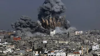 The image depicts a scene of destruction with a large explosion occurring in an urban area. Thick, dark gray smoke billows into the sky, rising above the buildings in the foreground. The skyline is dominated by closely packed structures, showing signs of damage. The overall atmosphere conveys a sense of chaos and devastation, characteristic of an area affected by conflict or military action.