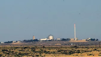 The image depicts a desert landscape with a prominent facility in the background. This facility features a large, domed structure, likely indicative of a scientific or research establishment, alongside tall smokestacks or cooling towers. The setting appears arid, with sparse vegetation, and the horizon stretches into the distance, emphasizing the isolation of the facility. The overall atmosphere conveys a sense of industrial or technological presence amidst a natural environment.
