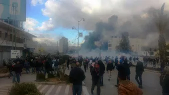 L'image montre une scène de manifestation dans une ville, avec une foule rassemblée dans une rue. Il y a des nuages de fumée et des signes de tension, suggérant des troubles ou des affrontements. Les gens marchent, certains semblent fuir la fumée, tandis que des bâtiments et des arbres sont visibles en arrière-plan. Le ciel est partiellement nuageux, ajoutant à l'atmosphère dramatique de la scène.