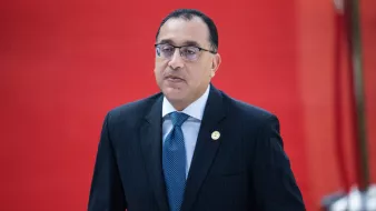 The image features a man dressed in a formal dark suit with a light blue tie. He appears to be in a professional setting, as suggested by the suit and tie. The background is bright red, which adds a striking contrast to his attire. The man looks serious and focused, possibly engaged in an official event or meeting.