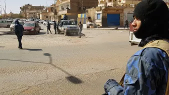 تظهر الصورة مشهداً في منطقة حضرية، حيث يوجد عدد من رجال الأمن يرتدون زيًّا رسميًّا وقبعات سُوداء. أحدهم يحمل سلاحًا ويقف في المقدمة، بينما يتواجد آخرون في الخلفية. تشهد المنطقة حركة مرورية، مع وجود سيارات متوقفة في الشارع. المباني المحيطة تظهر بألوان متنوعة، وبعضها يبدو أنه مغلق. تعكس الصورة أجواءً من اليقظة والحذر الأمني.