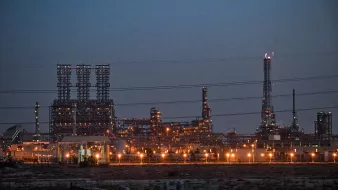 The image depicts an industrial facility, likely a refinery or manufacturing plant, illuminated by numerous lights during dusk. The skyline features tall structures, including towers and stacks, with a complex arrangement of pipes and machinery. The surrounding area appears barren, with some electrical towers and vegetation visible in the foreground. The overall atmosphere suggests a busy industrial site functioning into the evening.