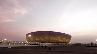 L'image montre un grand stade moderne à l'architecture distinctive, situé sous un ciel teinté de nuances pastel au coucher du soleil. Le stade possède une structure en forme d'ellipse avec une façade brillante, peut-être en métal ou en verre, qui capte la lumière ambiante. On aperçoit quelques personnes marchant devant le stade, soulignant sa taille imposante. L'ensemble de la scène dégage une ambiance calme et majestueuse.