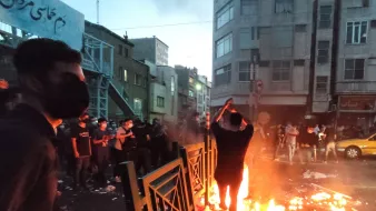 در این تصویر، گروهی از افراد در خیابانی در حال تظاهرات هستند. در مرکز عکس، فردی در حال آتش زدن شیئی است که آتش به‌طور واضحی شعله‌ور شده است. دور تا دور او زباله‌ها و آشغال‌ها پراکنده شده و برخی از معترضان در حال تماشا هستند. بیشتر افراد ماسک به‌دهن دارند و فضای عمومی به نظر شلوغ و تنش‌زا می‌رسد. ساختمان‌های اطراف و نور پایین آسمان نیز به حالت دراماتیک تصویر افزوده است.