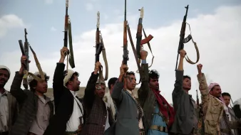 The image shows a group of men holding up rifles in the air. They are standing in a line, with some raising their right arms in a fist while others are focusing on the guns. The setting appears to be outdoors, with a cloudy sky in the background. The men are wearing traditional attire, which varies among them, and there is an overall sense of solidarity and determination in their postures.