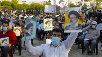 در تصویر، جمعیتی از افراد در یک فضای باز دیده می‌شود که روی صندلی نشسته‌اند و تعدادی از آن‌ها در حال نگه‌داشتن پوسترهایی از یک شخصیت سیاسی هستند. مردم ماسک به‌صورت دارند و در پس‌زمینه، تعدادی پرچم و تابلوهای نمایشی نیز وجود دارد. این جمعیت به نظر می‌رسد که در یک رویداد یا حمایت سیاسی شرکت کرده‌اند.