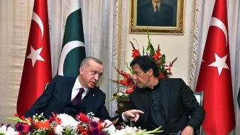 The image features two men engaged in conversation, likely discussing matters of significance. They are seated at a table adorned with flowers, and the backdrop includes flags of Turkey and Pakistan prominently displayed. A portrait of a historical figure hangs on the wall above them. Both individuals appear to be in formal attire, suggesting an official or diplomatic context for their meeting.