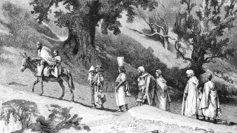 L'image représente une scène en noir et blanc où plusieurs personnages se déplacent sur un chemin. Au premier plan, un homme est assis sur un âne, tandis qu'un autre homme le guide à pied. D'autres figures, habillées de vêtements traditionnels, marchent derrière eux. La végétation environnante est constituée d'arbres, certains dépouillés, suggérant une ambiance peut-être désertique ou rural. L'illustration dégage une atmosphère historique, typique des gravures anciennes.