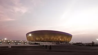 تظهر الصورة ملعبًا حديثًا بتصميم جذاب، يتميز بواجهة ذهبية وأضواء تضيء محيطه. السماء خلف الملعب تأخذ ألوانًا دافئة، بينما يظهر عدد من الأشخاص يمشون في المنطقة المحيطة. يبدو الملعب كبيرًا ومهيبًا، مما يعكس الهندسة المعمارية المعاصرة.