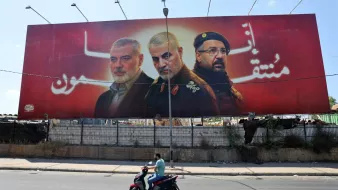 L'image montre une grande affiche publicitaire avec trois portraits d'hommes, probablement des figures militaires ou politiques, sur fond rouge. Un homme à moto passe devant cette affiche, qui semble être placée le long d'une route. L'environnement semble urbain, avec des déchets visibles au fond.