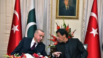 En la imagen se pueden ver a dos hombres conversando en un ambiente formal, rodeados de banderas de Turquía y Pakistán. Hay un arreglo floral colorido en la mesa, y detrás de ellos se encuentra un retrato en la pared. La escena transmite un ambiente de diplomacia y colaboración entre los dos países.