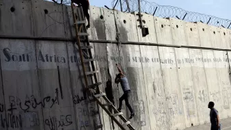 L'image montre une scène où deux personnes tentent de monter ou de descendre d'un mur de béton. Une échelle est appuyée contre le mur, et il y a des barbelés au sommet. Le mur présente également des graffitis ou des inscriptions, et l'environnement semble urbain. Les personnes semblent concentrées sur leur tâche, soulignant une dynamique d'effort et de défi.