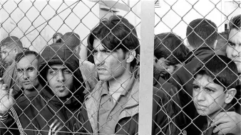 L'image montre un groupe d'hommes, visiblement en attente, qui se trouvent derrière une clôture en fil de fer. Leur expression faciale révèle une certaine anxiété et tension. Ils semblent chercher à voir ou à communiquer avec le monde extérieur. Le cadre en noir et blanc accentue l'intensité de leurs émotions et la gravité de la situation.