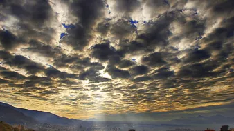 Cette image montre un paysage urbain avec des bâtiments s'étendant dans la vallée, sous un ciel dramatique rempli de nuages. Les nuages sont denses et variés, créant une texture visuelle intéressante, tandis que des rayons de lumière percent à travers eux, illuminant partiellement la ville en contrebas. On peut également apercevoir des montagnes à l'arrière-plan, ajoutant une dimension naturelle à la scène. L'atmosphère est à la fois majestueuse et sereine, capturant un moment poétique de la journée.
