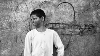 L'image montre un garçon qui se tient contre un mur texturé, où l'on peut voir des éraflures et des graffitis. Il porte un t-shirt blanc légèrement sale et un pantalon beige. Son expression semble pensive ou mélancolique. Le décor derrière lui, avec un dessin de cœur, contraste avec son attitude, créant une atmosphère intéressante. Cela donne une impression de nostalgie ou de réflexion sur son environnement.