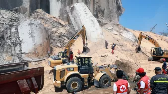 L'image montre un site de construction ou de déblaiement où des engins de chantier, tels que des pelles mécaniques et des bulldozers, sont en action. Au premier plan, des travailleurs en uniforme, y compris des membres de la Croix-Rouge, supervisent les opérations. En arrière-plan, on peut voir des débris et des structures effondrées, suggérant une situation d'urgence ou de nettoyage après un incident. Le ciel est dégagé et ensoleillé, ajoutant un contraste avec l'environnement de travail.