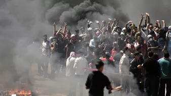 L'image montre un groupe de personnes rassemblées dans une atmosphère tendue. Elles sont entourées de fumée noire, probablement causée par des feux. Certains manifestants lèvent les bras en signe de protestation. On peut sentir une ambiance de colère et de résistance, avec quelques personnes qui semblent brandir des objets. Le décor est chaotique, évoquant une situation de crise ou de manifestation.