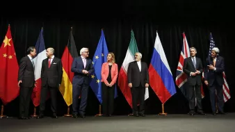 L'image montre une scène officielle avec plusieurs personnes debout en rang, chacune représentant un pays ou une organisation distincte. Derrière eux, il y a des drapeaux de différents pays, notamment la Chine, l'Allemagne, les États-Unis, la Russie et l'Union européenne, entre autres. Les participants semblent engagés dans une discussion ou une négociation, chacun portant un costume formel. L'atmosphère est sérieuse, probablement liée à des pourparlers diplomatiques ou à un sommet international.