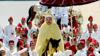 L'image montre un roi entouré de nombreux participants, tous vêtus de vêtements traditionnels. Le roi est monté sur un cheval, revêtu d'une cape jaune et portant un chapeau distinctive. Au-dessus de lui se trouve un parasol rouge. Les membres de l'entourage portent des turbans rouges, tandis que d'autres sont habillés en blanc. L'arrière-plan semble être une cérémonie officielle, avec des spectateurs et un décor sobre. Cette scène évoque une ambiance de tradition et de solennité.