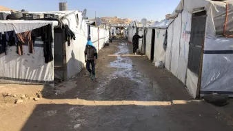 The image depicts a narrow path between rows of temporary tents or makeshift shelters, likely in a refugee or displaced persons camp. The tents are primarily white and appear to be made from tarpaulin or similar materials. There are clothes hanging outside some of the tents, suggesting attempts to dry them. The ground is muddy, indicating recent rain, and the surrounding area appears arid with few visible structures beyond the camp. The scene conveys a sense of modest living conditions and the challenges faced by individuals in such environments.