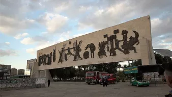 L'image montre une grande structure en béton surmontée de silhouettes découpées dans le matériau, représentant des figures humaines et des scènes de la vie quotidienne. Cette œuvre d'art moderne semble se dresser dans un espace public, entourée de véhicules et de personnes. Le ciel est nuageux, suggérant une ambiance calme et une lumière douce. En arrière-plan, on peut voir des bâtiments et des éléments d'urbanisme typiques d'une ville. L'ensemble évoque un mélange de culture et d'art, tout en étant intégré dans la vie citadine.
