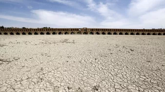 L'image montre un paysage aride avec des sols craquelés, indiquant une sécheresse sévère. À l'arrière-plan, on aperçoit un grand pont historique avec plusieurs arches. Le ciel est dégagé avec quelques nuages, et l'absence d'eau dans la zone suggère des conditions environnementales préoccupantes. Cette scène illustre les effets de la déshydratation sur un espace habituellement aquatique.