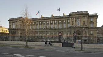 The image depicts a grand building with a classical architectural style, likely a government or official structure. The facade features ornate details, including large windows and decorative elements. French flags are visible atop the building, indicating its significance. There is a wrought iron fence in front, along with columns that enhance the structure's elegance. The surrounding area appears to be urban, with a street visible in the foreground, suggesting the building is located in a city. The overall scene conveys a sense of history and importance.