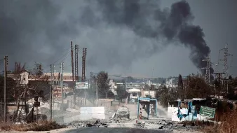 The image depicts a desolate street under a dark, smoke-filled sky. In the background, billowing plumes of black smoke suggest an ongoing fire or explosion. The foreground shows a damaged road with debris, potentially hinting at a conflict zone. There are signs of destruction, including burnt vehicles and abandoned structures, contributing to an overall atmosphere of turmoil and devastation. The landscape appears barren with a few power lines and distant buildings, reflecting the impact of violence on the area.