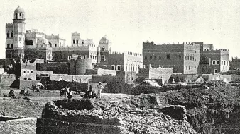 L'image présente un paysage urbain ancien, probablement d'une ville historique. On peut voir des bâtiments en pierre avec des détails architecturaux variés, tels que des tours et des terrasses. À l'avant-plan, on aperçoit des travailleurs, probablement engagés dans des activités agricoles ou de construction. La scène évoque une atmosphère à la fois rustique et animée, avec un ciel clair et des montagnes en arrière-plan qui ajoutent à la dimension historique et géographique de l'endroit. L'ensemble dégage une impression de temps révolu et de tradition.