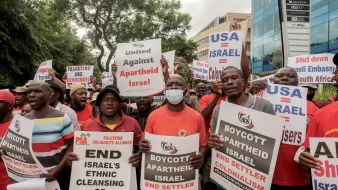L'immagine ritrae un gruppo di manifestanti impegnati in una protesta. I partecipanti, vestiti in gran parte con magliette rosse, tengono in mano cartelli con testi che chiedono la fine dell'apartheid in Israele, il boicottaggio delle politiche israeliane e l'unità contro il colonialismo. Alcuni cartelli menzionano specificamente il "Pulizia etnica di Gerusalemme" e fanno riferimento ad azioni di protesta contro l'ambasciata degli Stati Uniti in relazione a questi temi. L'atmosfera sembra carica di energia e determinazione.