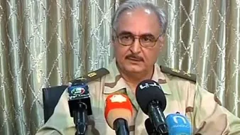 L'image montre un homme portant un uniforme militaire avec des rangs. Il est assis devant plusieurs microphones, suggérant qu'il est en train de donner une déclaration ou une interview. L'arrière-plan est composé de rideaux clairs, et l'homme a une expression sérieuse. On sent qu'il s'agit d'une situation officielle.