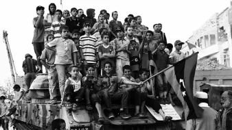 L'image montre un grand groupe d'enfants rassemblés sur un char, probablement dans un contexte de célébration ou de manifestation. Les enfants affichent diverses expressions, allant de la joie à la curiosité. Ils portent des vêtements colorés, souvent rayés, et certains tiennent un drapeau. En arrière-plan, on peut voir des bâtiments et des ruines, suggérant un environnement marqué par des conflits ou des événements historiques. L'image est en noir et blanc, ce qui accentue le côté dramatique de la scène.