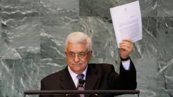 L'image montre un homme debout devant un pupitre de l'Organisation des Nations Unies (ONU). Il porte un costume noir avec une cravate et des lunettes. L'homme soulève un document, probablement un discours ou une déclaration, en s'adressant à l'assemblée. L'arrière-plan est décoré de panneaux en pierre, typiques des salles de conférence de l'ONU.