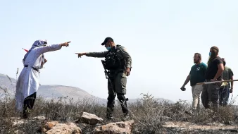 L'image montre une scène de tension entre un homme habillé dans un style traditionnel, tenant une sorte de bâton ou d'objet, qui semble pointer du doigt. Un policier ou un soldat, portant un uniforme et un gilet, est en position d'affrontement avec lui, pointant également vers l'homme. En arrière-plan, plusieurs autres personnes, visiblement des observateurs ou des participants à la scène, se tiennent. Le paysage environnant semble aride, avec des rochers. L'atmosphère est chargée et indique une situation potentiellement conflictuelle.