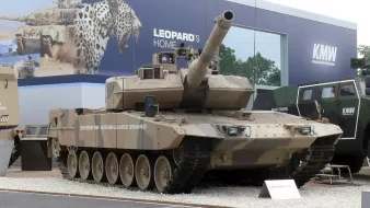 L'image montre un véhicule militaire, plus précisément un char de combat. Le char est peint dans un motif de camouflage beige et brun, typique pour se fondre dans des environnements désertiques. Il est équipé d'un canon long monté sur le dessus et possède plusieurs roues de chaque côté. En arrière-plan, on aperçoit des éléments d'exposition militaire, suggérant que le char est présenté lors d'un salon ou d'une exposition lié à la défense. Les détails tels que les plaques d'identification du char peuvent également être visibles.