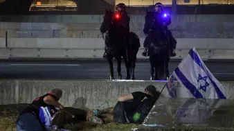 The image depicts a tense scene possibly related to a protest or civil unrest. In the foreground, two individuals appear to be on the ground, with one person lying down and another sitting nearby. They seem to be taking cover or seeking safety. In the background, two mounted police officers are visible, equipped with flashing lights on their helmets. There's also an Israeli flag positioned on the ground, suggesting that the situation might involve themes of national identity or political protest. The setting looks urban, with structures and concrete present, contributing to a sense of urgency in the atmosphere.