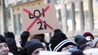 L'image montre un groupe de personnes manifestant. Au premier plan, une personne tient une pancarte en carton sur laquelle est écrit "LOI 21" avec une grande croix rouge qui la barre. Les participants portent des gants et des bonnets, suggérant qu'ils manifestent par temps frais. L'ambiance semble être celle d'une protestation, probablement contre la loi mentionnée.