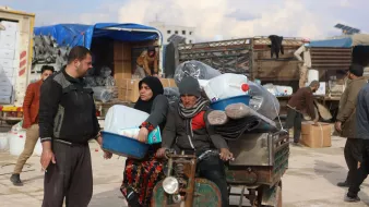 L'image montre une scène de marché ou de distribution dans un environnement rural. On y voit un homme et une femme discutant, tandis qu'un autre homme est assis sur un véhicule à trois roues, probablement en train de transporter des marchandises. Le décor entoure des containers en plastique, et plusieurs personnes s'affairent à décharger ou à déplacer des articles. Le ciel est nuageux, suggérant une atmosphère d'activité et d'échange.