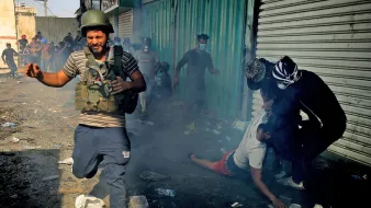 تظهر الصورة مشهدًا من الفوضى، حيث يركض رجل يرتدي خوذة ودروعًا واقية وسط دخان كثيف في منطقة يبدو أنها تعرضت لأعمال شغب أو احتكاك. في الخلفية، يظهر عدد من الأشخاص، يبدو أن بعضهم يتفاعل بشكل عاطفي مع الوضع. الأرض مليئة بالقمامة، مما يعكس حالة من الاضطراب والانزعاج. الجو العام يوحي بالتوتر والخطر.