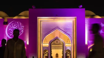 L'image montre une grande structure architecturale éclairée de manière colorée, principalement en violet. On peut voir un portail monumental avec des motifs décoratifs, typiques de l'architecture islamique. À l'avant, des silhouettes de personnes marchent, créant une atmosphère vivante et dynamique. Au fond, des éléments de design comme des arcs et des motifs sont visibles, ajoutant une touche d'élégance à l'ensemble de la scène. L'éclairage nocturne met en valeur les détails architecturaux et crée une ambiance fascinante.