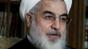 الصورة تظهر رجلًا يرتدي عمامة بيضاء ويبدو أنه يتحدث بحماس. لديه لحية بيضاء ويرتدي زيًا تقليديًا. يظهر الشخص في مكان مغلق مع خلفية تحتوي على رفوف كتب. تعكس تعابير وجهه اهتمامًا ونشاطًا أثناء الحوار.