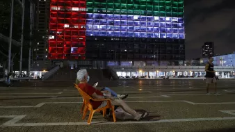 L'image montre une scène nocturne dans une ville. Au premier plan, un homme âgé est assis sur une chaise orange, en train de regarder son téléphone. En arrière-plan, un bâtiment imposant est illuminé avec des lumières de différentes couleurs, formant un motif qui ressemble à un drapeau. Les couleurs rouge, vert et bleu s'alternent sur la façade du bâtiment, créant une ambiance vibrante. D'autres personnes se déplacent dans l'espace public, ajoutant de la vie à la scène.