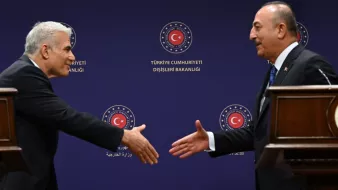 في الصورة، يظهر اثنان من الرجال يقفان في مواجهة بعضهما البعض. يبدو أن أحدهما يقدم يده لمصافحة الآخر. خلفهما يظهر علم تركيا مع شعار وزارة الخارجية التركية. الأجواء تبدو رسمية، مما يشير إلى أن اللقاء قد يكون متعلقاً بموضوع دبلوماسي أو رسمي بين البلدين.