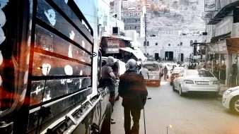 L'image montre une rue animée avec des véhicules garés et des passants. On aperçoit un homme âgé marchant avec une canne, tandis que d'autres personnes se déplacent. À l'arrière-plan, des bâtiments et des collines sont visibles, suggérant un environnement urbain. La photo semble avoir un effet de noir et blanc, avec des touches de couleur chaude, créant une ambiance nostalgique.