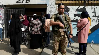 تظهر في الصورة مجموعة من الأشخاص يتجمعون في مكان يبدو أنه مدخل لمبنى، حيث يقف جندي مسلح أمام المدخل. يرتدي الجندي زيًا عسكريًا ويحمل سلاحًا، بينما تتنوع ملابس الأشخاص الذين حوله، حيث ترتدي بعض النساء العباءات والحجاب. تعكس الصورة أجواء من الحذر والاحتياط، مع وجود جندي للحماية أو تنظيم الصفوف.