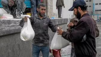 L'immagine mostra un gruppo di uomini che stanno ricevendo o scambiando sacchetti. Gli uomini sembrano essere in un ambiente di lavoro, con segni di polvere sui loro vestiti, suggerendo che stiano partecipando a un'attività che richiede impegno fisico. I sacchetti possono contenere materiali o forniture, e l'atmosfera appare focalizzata e praticante. Sullo sfondo ci sono altri uomini e alcune strutture, che contribuiscono a un contesto di lavoro collettivo.
