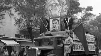L'image représente un défilé historique avec un camion décoré. Sur le camion, on peut voir des portraits de figures politiques, probablement des leaders communistes, entourés de drapeaux. Un homme en uniforme se tient debout sur le camion, tandis que d'autres personnes défilent en arrière-plan. L'atmosphère semble festive et engagée, typique des manifestations politiques d'époque. Les bâtiments et les passants en arrière-plan ajoutent un contexte urbain à la scène.